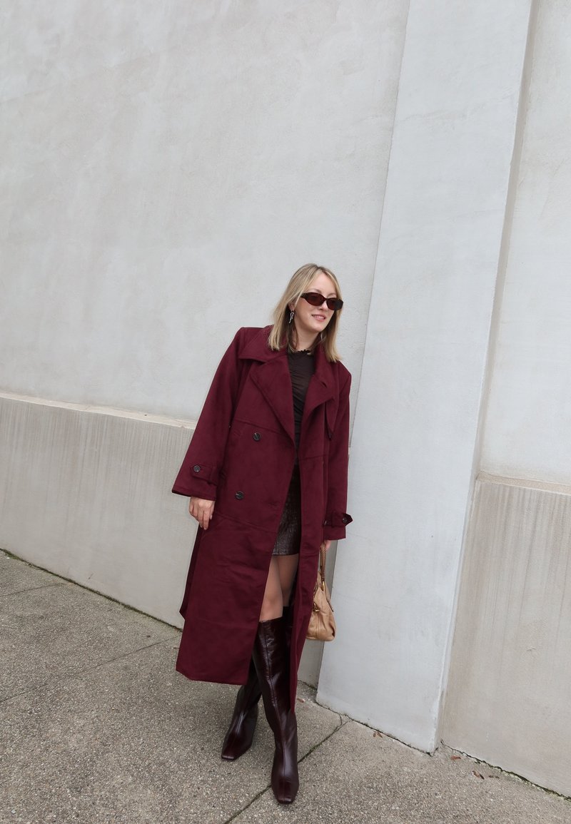 Trench-coat bordeaux avec des boutons croisés et des larges revers, assorti à un haut ajusté foncé, une mini-jupe et des bottes montantes à bout carré.