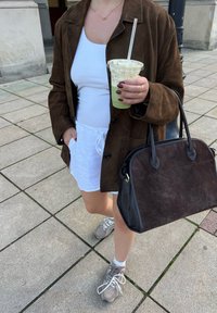 Barna szarvasbőr túlméretezett dzseki, fehér, zsinórral ellátott rövidnadrág, szürke sportcipő, fekete kézitáska, átlátszó pohár zöld itallal, semleges színű járda háttér.