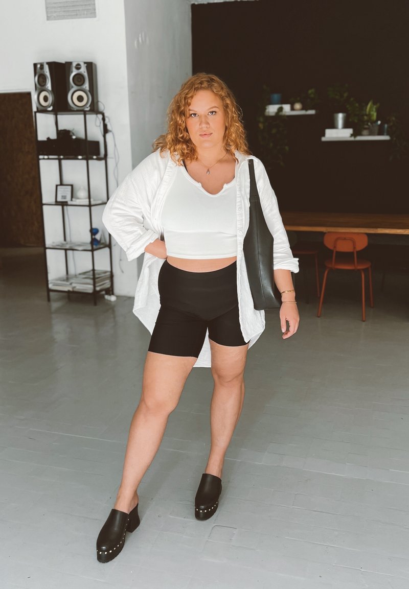 Λευκό oversized πουκάμισο, εφαρμοστό λευκό crop top, μαύρες σορτς ποδηλασίας, και μαύρες ξηρές παντόφλες με τρουκς. Φωτεινός ξύλινος και πράσινος διακόσμος στο παρασκήνιο.