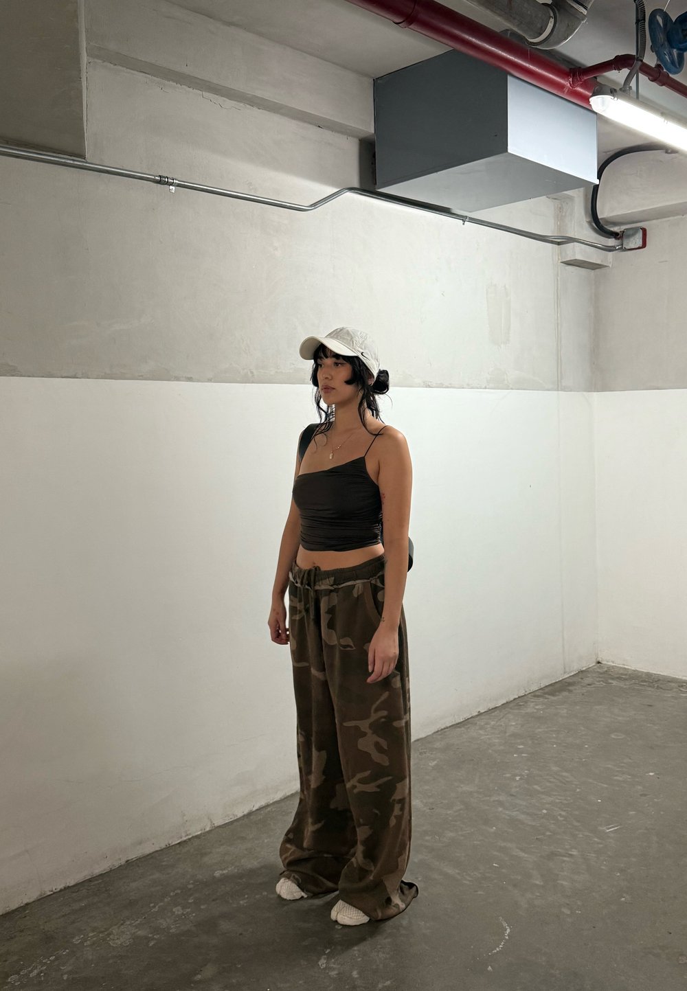 Vrouw met zwart haar, gekleed in een witte pet, een zwart crop top en losse camouflagebroek, staat in een minimalistisch industrieel ingerichte ruimte met een betonnen vloer.