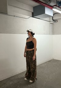 Vrouw met zwart haar, gekleed in een witte pet, een zwart crop top en losse camouflagebroek, staat in een minimalistisch industrieel ingerichte ruimte met een betonnen vloer.