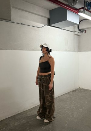Mujer con cabello negro que lleva una gorra blanca, un top corto negro y pantalones holgados de camuflaje, de pie en una habitación minimalista de estilo industrial con suelo de hormigón.