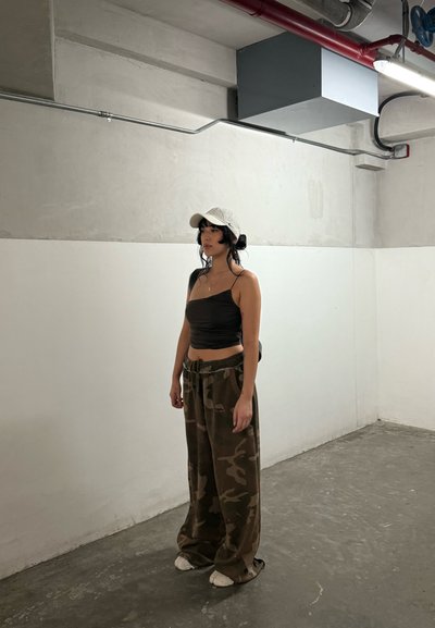 Mujer con cabello negro que lleva una gorra blanca, un top corto negro y pantalones holgados de camuflaje, de pie en una habitación minimalista de estilo industrial con suelo de hormigón.