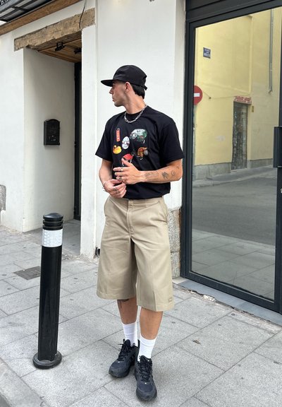 Camiseta negra con gráficos, pantalones cortos beige y zapatillas negras. Los accesorios incluyen una cadena de plata y pulseras. Entorno urbano con acera.