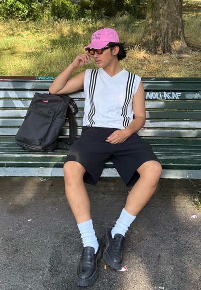 Top blanco sin mangas con rayas negras, pantalones cortos negros, mocasines negros, calcetas blancas, gorra rosa y una mochila negra sobre un banco verde.