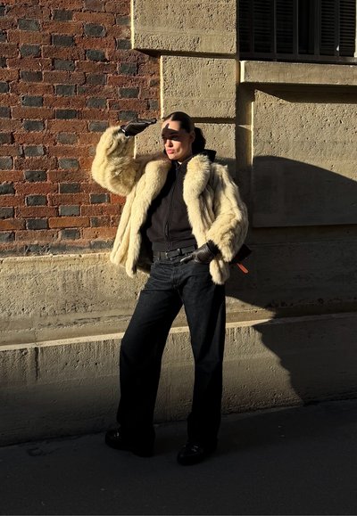 Persona con abrigo de piel, guantes negros y pantalones oscuros, cubriéndose los ojos mientras está de pie contra una pared de ladrillo y piedra iluminada por el sol.