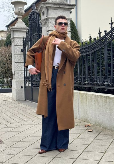 Hombre con abrigo largo marrón y bufanda, pantalones anchos oscuros, gafas de sol, zapatos marrones, llevando un bolso de cuero marrón, de pie junto a una puerta de hierro forjado.