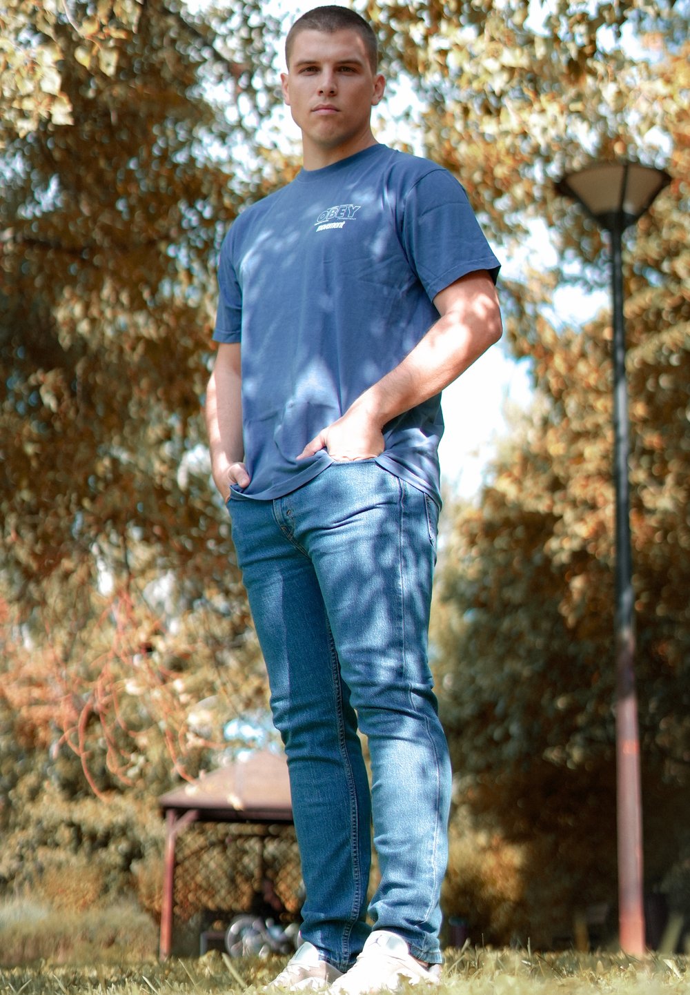 Blå bomulds t-shirt med korte ærmer, lyseblå jeans og hvide sneakers. Stående i en park med træer og en lygtepæl i baggrunden.