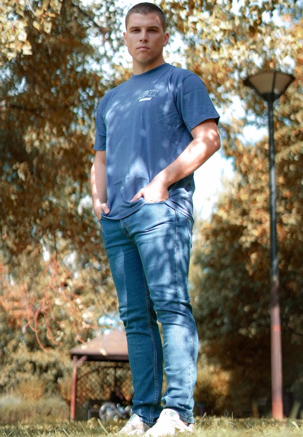 Blå bomulds t-shirt med korte ærmer, lyseblå jeans og hvide sneakers. Stående i en park med træer og en lygtepæl i baggrunden.