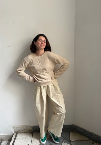 Mujer con cabello corto y oscuro está apoyada contra una pared blanca, ojos cerrados, manos en las caderas, lleva suéter beige, pantalones anchos y zapatos verdes a cuadros.