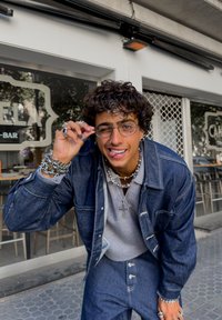 Young man with curly hair wearing glasses, denim jacket, and layered necklaces, adjusting glasses and smiling outside a café.