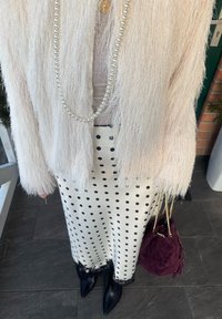 Femme portant une veste beige duveteuse, un collier de perles, une jupe blanche à pois noirs, des bottes noires, et tenant un sac à main en daim violet.