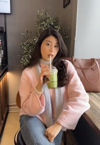 Mujer con un suéter rosa y una bufanda gris bebiendo un latte de matcha helado mientras está sentada en una mesa de madera con un bolso marrón cerca.