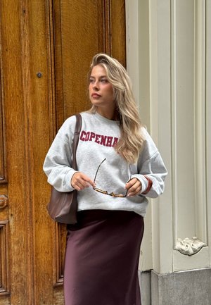 Sudadera en gris con "COPENHAGUE" en rojo oscuro, combinada con una falda burdeos. Bolsa marrón, sosteniendo gafas de sol, contra un fondo de madera.