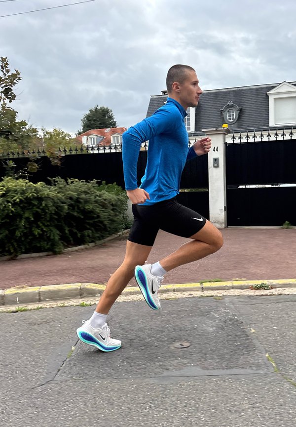 Un hombre con una camiseta azul y pantalones cortos negros corre por una calle residencial, pasando junto a una casa con verja bajo un cielo nublado.