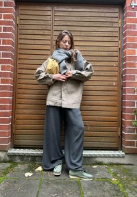Beige oversized jakke, grå tørklæde, brede grå bukser og lysegrønne sko. Står foran en dør med trælameller og en murstensvæg.