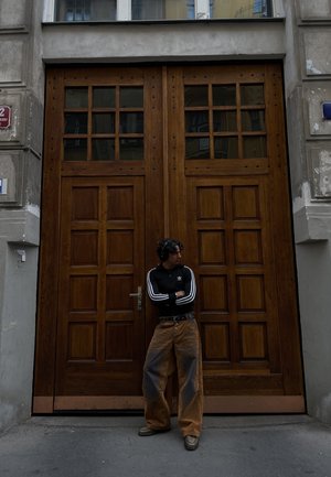 Chaqueta negra con cremallera y rayas blancas, pantalones de pierna ancha color beige, y auriculares, de pie frente a grandes puertas dobles de madera con paneles de vidrio.