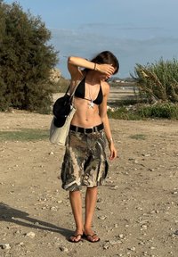 Črn bikini vrh, cargo kratke hlače s kamuflažnim vzorcem, sandali, srebrna ogrlica s pentljo v obliki zvezde, nosi svetlo obarvano torbico.