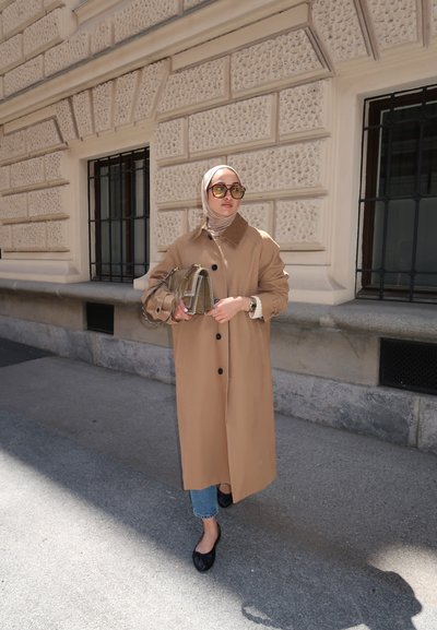 Trench beige con una parte frontal abotonada, mangas largas y un cuello; usado sobre unos jeans azul claro y bailarinas negras. Accesorio: bolso pequeño con estampado.