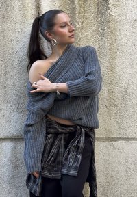 Femme avec une queue de cheval haute portant un pull gris à épaules dénudées, des boucles d'oreilles anneaux, une chemise à carreaux nouée à la taille et un pantalon noir, posant devant un mur texturé.