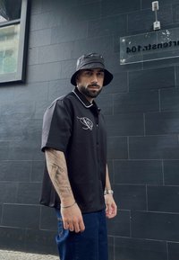 Črna kratka srajca s kratkimi rokavi in gumbi, z belim obrobo in grafičnim načrtom, v kombinaciji s temno modrimi jeans hlačami in črnim kendijem.