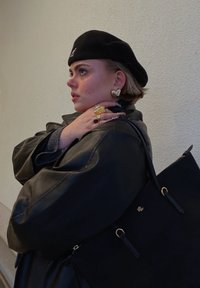 Μαύρο δερμάτινο oversized μπουφάν, μαύρο μπερεδάκι, χρυσαφένια σκουλαρίκια σε σχήμα καρδιάς, πολλαπλοί δακτύλιοι και μια μαύρη τσάντα tote με χρυσές λεπτομέρειες.