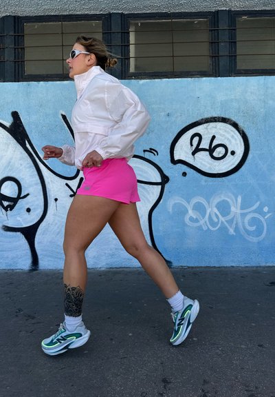 Mujer corriendo con chaqueta blanca, pantalones cortos rosas y zapatillas verdes junto a una pared azul con grafiti en blanco y negro y el número "26" en un bocadillo de diálogo.