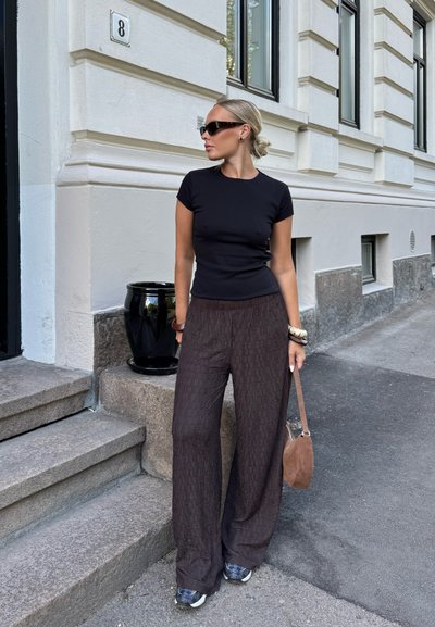 Mujer con cabello rubio recogido en un moño, lleva gafas de sol negras, camiseta negra ajustada, pantalones marrones anchos y zapatillas, de pie junto a unos escalones de piedra urbanos.