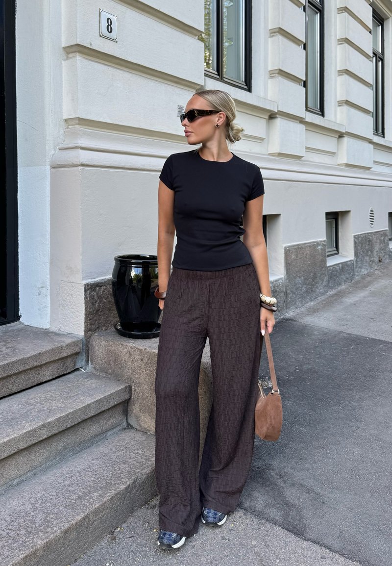 Femme aux cheveux blonds en chignon portant des lunettes de soleil noires, un t-shirt noir ajusté, un pantalon large marron et des baskets, debout près d'escaliers en pierre urbains.