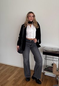 Zwarte blazer over een wit cropped blouseje met knopen en grijze jeans met wijde pijpen. Geaccentueerd met een zwarte tas en glanzende zwarte schoenen. Licht houten vloer.