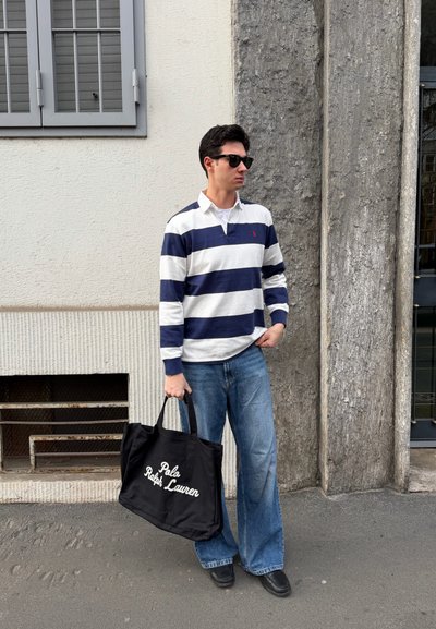 Hombre con gafas de sol, camiseta a rayas azul marino y blanca, vaqueros de pierna ancha, sosteniendo un bolso tote negro de Polo Ralph Lauren, de pie junto a una pared de piedra y blanca.