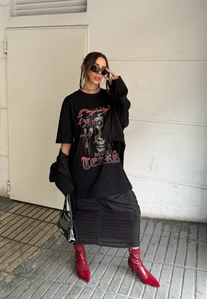 Camiseta negra de gran tamaño con estampado gráfico, superpuesta sobre una falda negra con pliegues. Botines rojos de punta y tobillo y un pequeño bolso negro. Gafas de sol negras.