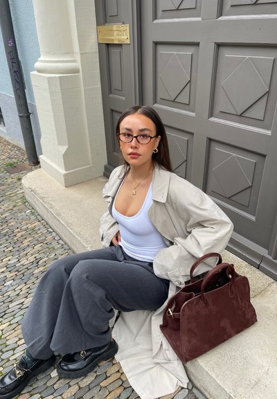 Trench beige sobre una camiseta blanca, pantalones anchos grises, zapatos negros y un bolso de ante rojo oscuro sobre una superficie de piedra.