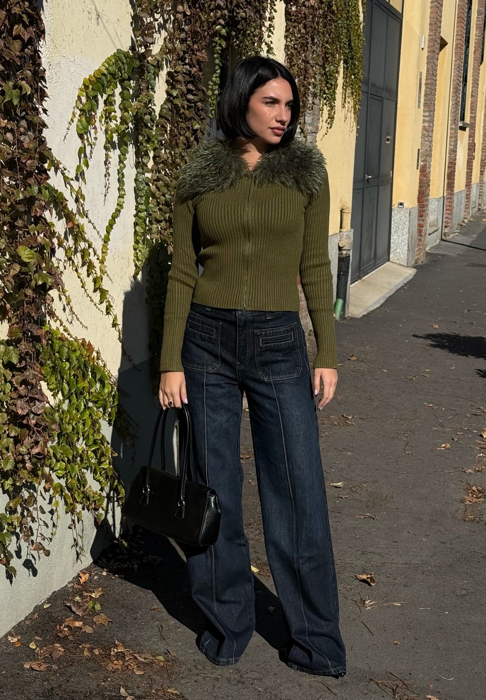 Olivgrüner gerippten Pullover mit Kunstfellkragen, kombiniert mit dunkelblauen weiten Jeans und einer schwarzen Handtasche. Vor einer mit Weinranken bewachsenen Wand.