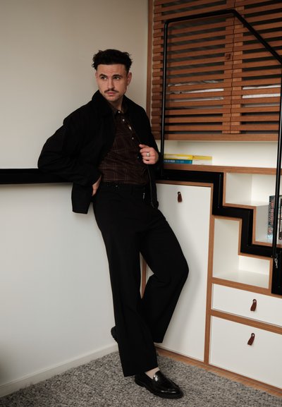 Hombre con bigote, vestido con chaqueta y pantalones oscuros, apoyado contra una pared blanca cerca de estanterías de madera con libros en una habitación interior.