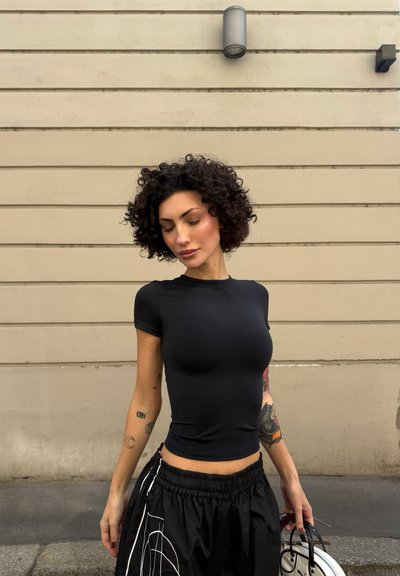 Mujer con el pelo rizado, vestida con una camisa negra ajustada y pantalones negros, de pie contra una pared beige sosteniendo un bolso blanco.