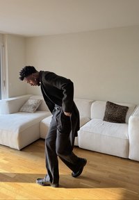 Homme en costume noir ajustant son pantalon dans un salon avec un canapé d'angle blanc et des coussins à motif animalier.