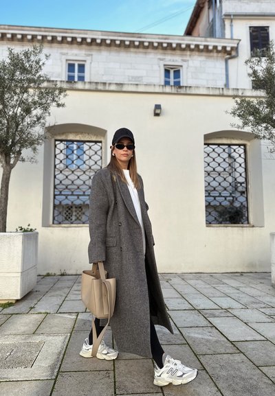 Abrigo de lana gris, camisa blanca, leggings negros, bolso beige y zapatillas blancas con acentos grises. El fondo presenta una pared de piedra y ventanas.