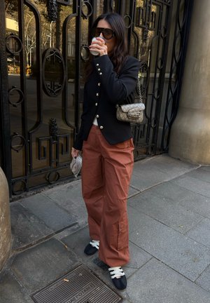 Mujer con blazer negro y pantalones color óxido bebe café estando junto a una puerta de metal negra ornamentada en la acera de la ciudad.