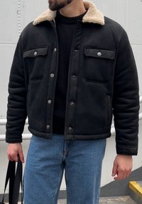 Veste noire matelassée avec un col en fausse fourrure crème, dotée de boutons-pression et de deux poches à rabat, portée sur un pull noir.