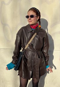 Manteau ceinturé en cuir marron avec grandes poches, fermeture à boutons et manches retroussées. Accessoires : un bandana rouge et des lunettes de soleil violettes.