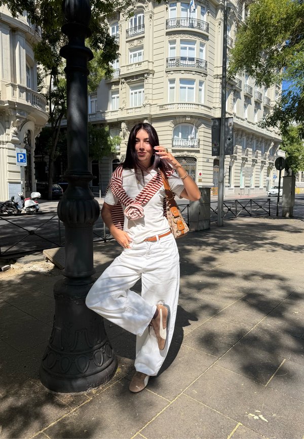 Jeune femme vêtue de vêtements blancs et d'un pull rayé noué autour des épaules, se tenant sur un trottoir ensoleillé de la ville près d'un lampadaire.