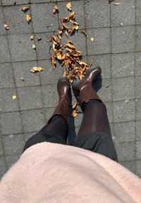 Brown leather knee-high boots with a textured finish, paired with black tights and a pink sweater, standing on a paved surface scattered with autumn leaves.