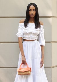Weißes, schulterfreies Crop-Top mit puffärmel, kombiniert mit einem gestuften weißen Maxikleid. Hält eine kleine orangefarbene und geflochtene Handtasche.