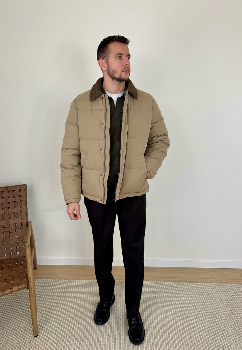 Homme portant une doudoune beige, un pull foncé, un pantalon noir et des chaussures noires, debout à l'intérieur près d'une chaise en osier sur un tapis clair.
