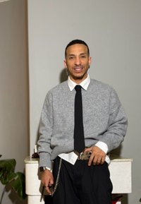 Silbergrauer Pullover, weißes Hemd mit Kragen, schwarze Krawatte und schwarze Hose. Auffälliger silberner Gürtel mit Kettendetail. Neutraler Hintergrund, grüne Pflanze in der Nähe.