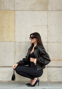 Mujer con top negro corto, chaqueta de cuero, leggings, tacones altos, gafas de sol grandes, sosteniendo un pequeño bolso negro, agachada contra una pared de piedra.