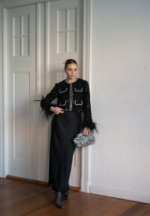 Chaqueta corta negra con lentejuelas y acentos de plumas, combinada con una falda negra de largo completo y un bolso de textura plateada.