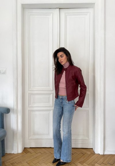 Mujer con cabello oscuro que lleva una chaqueta de cuero burdeos, un jersey de cuello alto rosa, vaqueros azul claro y zapatos negros, de pie frente a unas puertas dobles blancas cerradas.