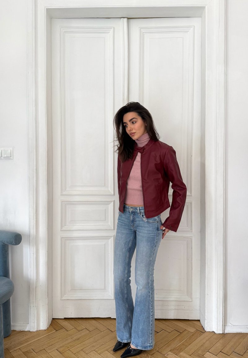 Ženska s temnimi lasmi, oblečena v bordo usnjeno jakno, roza ježko, svetlo modre kavbojke in čevlje črne barve, stoji pred zaprtimi belimi dvokrilnimi vrati.